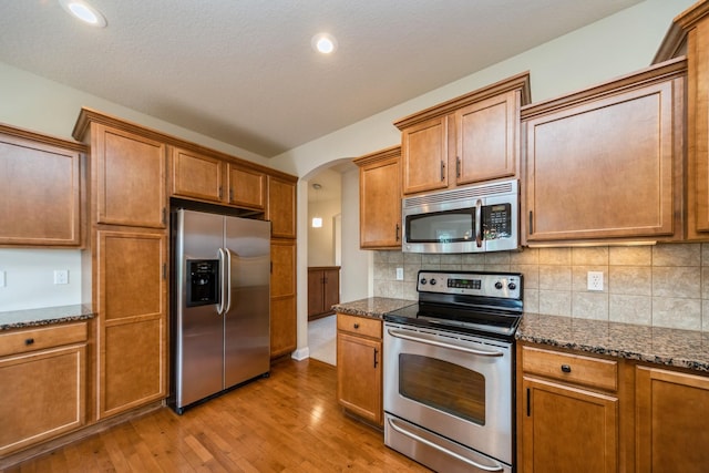 1420 Hannah Ln, Waukee, IA 50263 - kitchen featuring stainless steel appliances, arched walkways, light wood-type flooring, dark stone countertops, and brown cabinets
