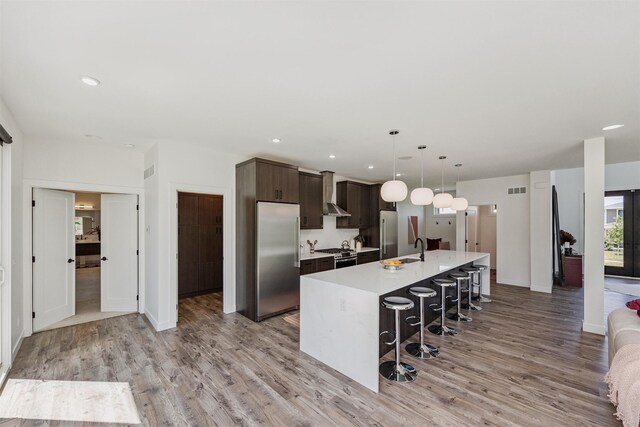 989 65th St, West Des Moines, IA 50266 - kitchen featuring stainless steel appliances, dark brown cabinets, an island with sink, light countertops, and wall chimney exhaust hood