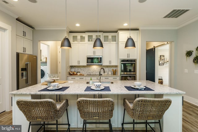 kitchen featuring appliances with stainless steel finishes, pendant lighting, washer / dryer, white cabinetry, and a kitchen island with sink