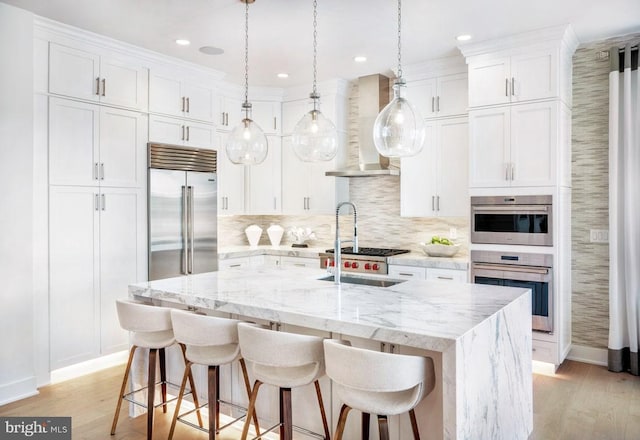 kitchen featuring backsplash, a kitchen island with sink, stainless steel appliances, light wood-type flooring, and wall chimney range hood