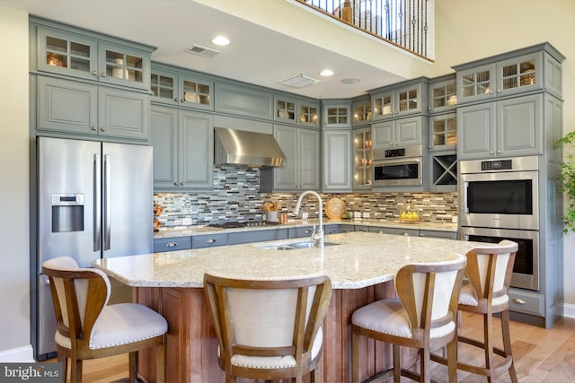 kitchen featuring an island with sink, appliances with stainless steel finishes, a breakfast bar area, light stone countertops, and wall chimney range hood