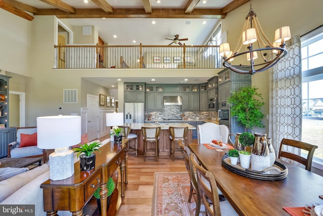 dining room with light hardwood / wood-style flooring, ceiling fan with notable chandelier, beam ceiling, and a high ceiling