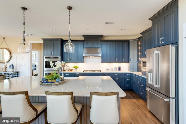 kitchen featuring a center island with sink, stainless steel appliances, decorative light fixtures, and light stone countertops