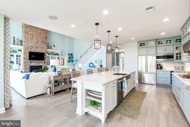 kitchen with a stone fireplace, built in appliances, a center island with sink, and light hardwood / wood-style floors