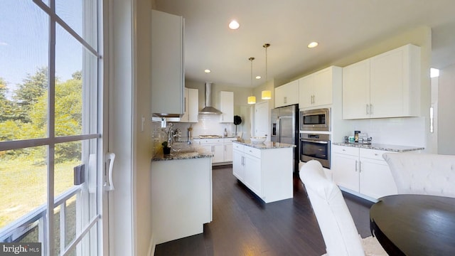 kitchen with appliances with stainless steel finishes, light stone counters, white cabinets, a kitchen island, and wall chimney exhaust hood