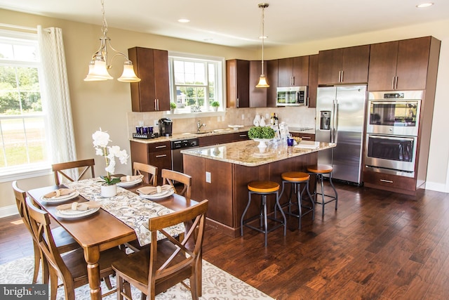 kitchen with appliances with stainless steel finishes, hanging light fixtures, a kitchen island, light stone counters, and dark hardwood / wood-style floors