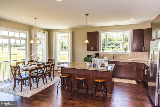 kitchen with light stone countertops, decorative light fixtures, stainless steel refrigerator, a kitchen island, and dark hardwood / wood-style flooring