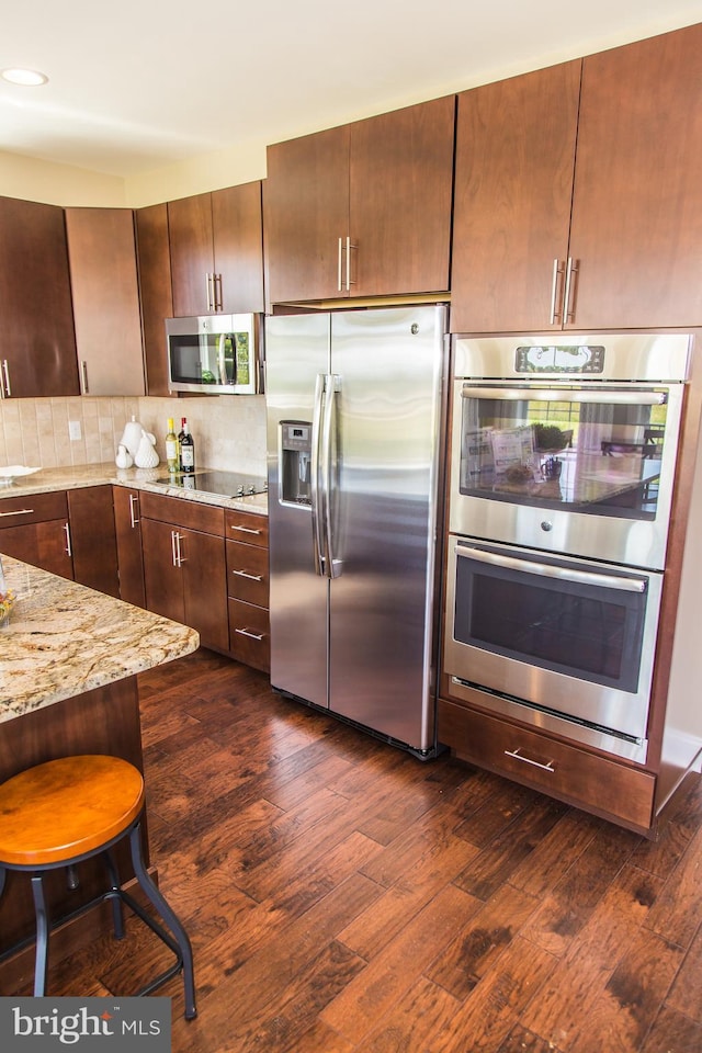 kitchen featuring dark wood-type flooring, a kitchen bar, stainless steel appliances, light stone countertops, and backsplash