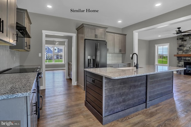 kitchen featuring ceiling fan, a center island with sink, a stone fireplace, backsplash, and dark hardwood / wood-style flooring