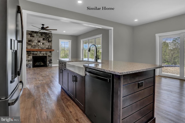 kitchen featuring ceiling fan, dark wood-type flooring, an island with sink, stainless steel appliances, and a stone fireplace