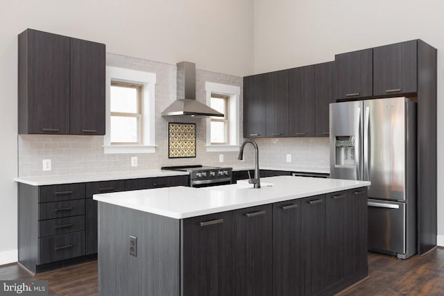 kitchen with dark wood-type flooring, wall chimney range hood, stainless steel appliances, and a kitchen island with sink