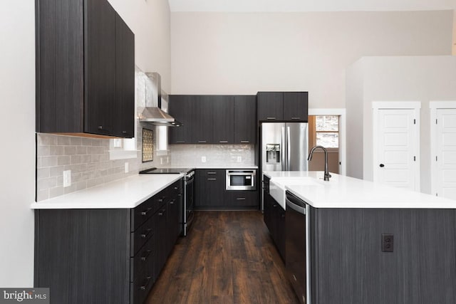 kitchen with dark hardwood / wood-style floors, backsplash, an island with sink, stainless steel appliances, and wall chimney range hood