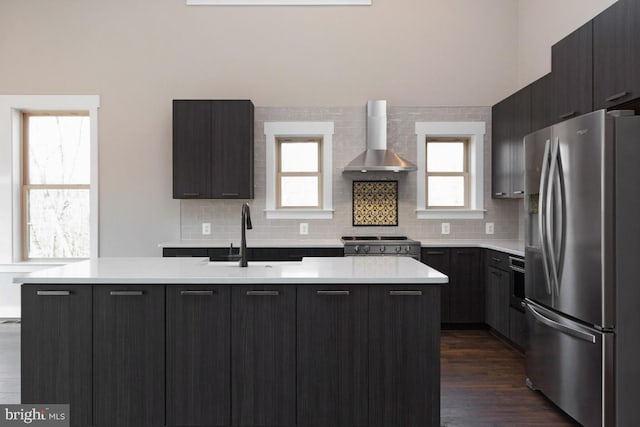 kitchen featuring appliances with stainless steel finishes, a kitchen island with sink, wall chimney range hood, and plenty of natural light