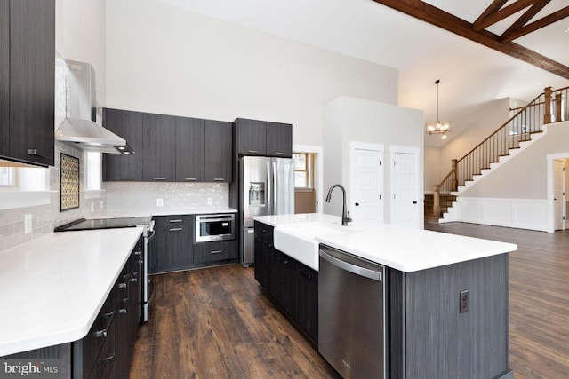 kitchen featuring wall chimney range hood, dark wood-type flooring, stainless steel appliances, a chandelier, and a kitchen island with sink