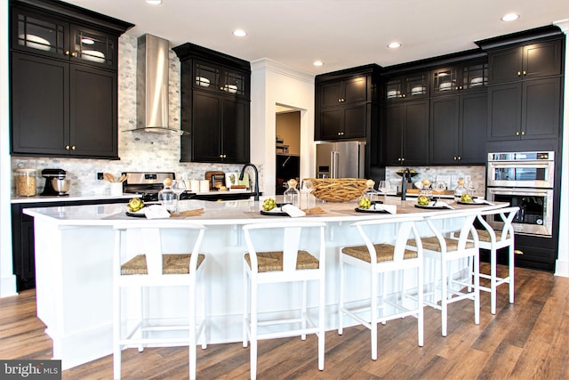 kitchen with wall chimney range hood, a kitchen breakfast bar, a center island with sink, and appliances with stainless steel finishes