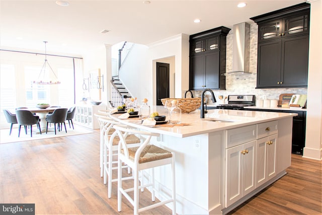 kitchen featuring hanging light fixtures, light wood-type flooring, wall chimney range hood, tasteful backsplash, and a kitchen island with sink