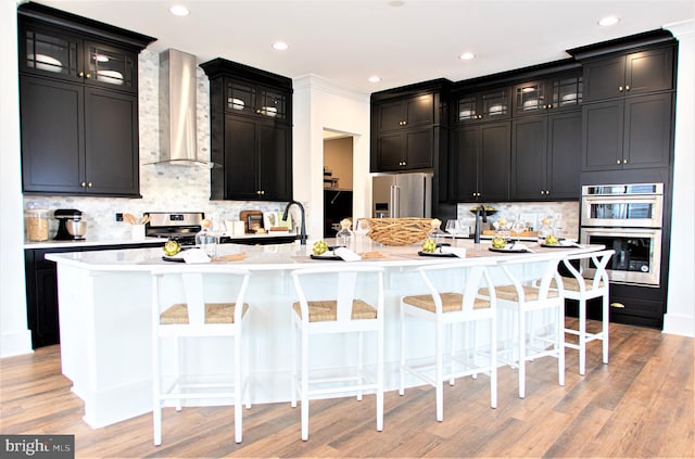 kitchen with wall chimney range hood, an island with sink, a kitchen bar, and stainless steel appliances