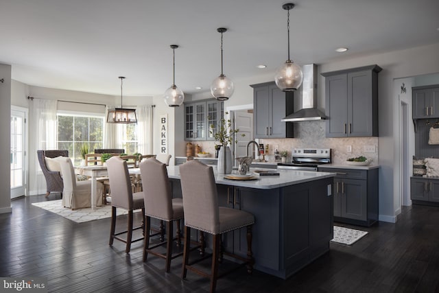 kitchen featuring an island with sink, decorative light fixtures, wall chimney exhaust hood, dark wood-type flooring, and electric stove