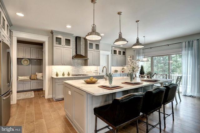 kitchen with wall chimney exhaust hood, stainless steel appliances, hanging light fixtures, and an island with sink