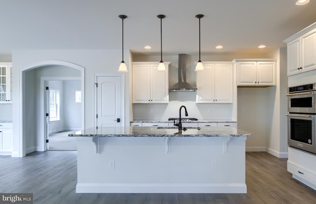 kitchen with dark stone countertops, white cabinets, an island with sink, and wall chimney exhaust hood