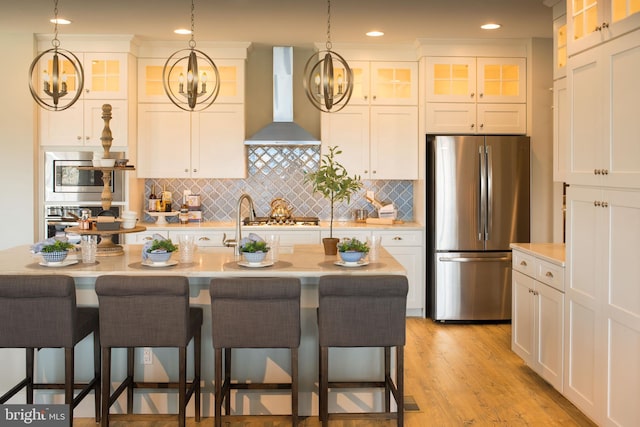 kitchen with wall chimney range hood, appliances with stainless steel finishes, white cabinetry, a center island with sink, and decorative backsplash