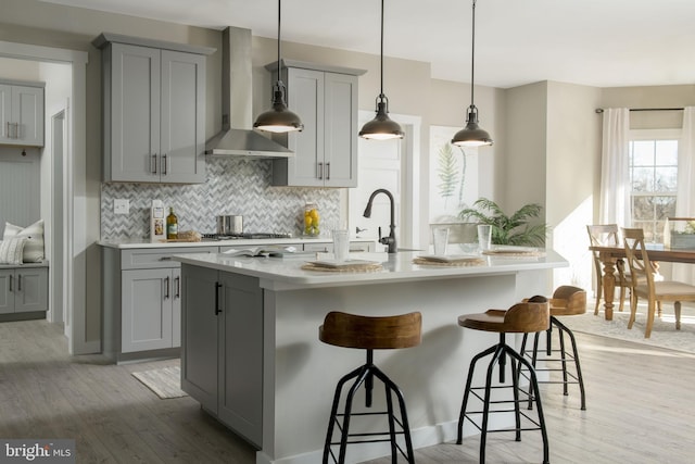 kitchen featuring a center island with sink, gray cabinets, wall chimney exhaust hood, and light hardwood / wood-style floors