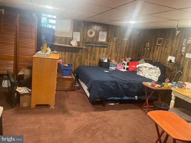 bedroom with a paneled ceiling, dark colored carpet, and wood walls