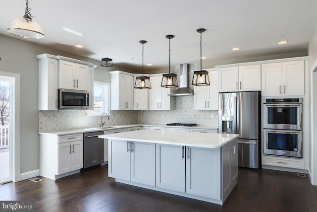 kitchen with wall chimney range hood, hanging light fixtures, appliances with stainless steel finishes, and dark wood-type flooring