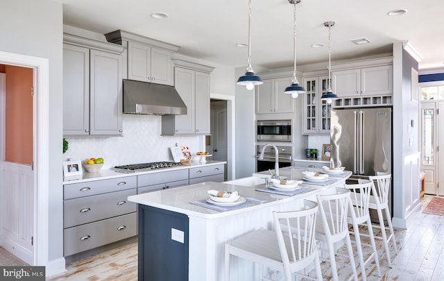 kitchen featuring an island with sink, built in appliances, hanging light fixtures, light hardwood / wood-style flooring, and a breakfast bar area