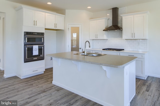 kitchen featuring sink, wall chimney range hood, multiple ovens, and a kitchen island with sink