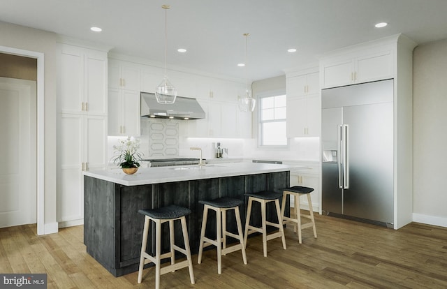 kitchen with light hardwood / wood-style floors, an island with sink, stainless steel built in fridge, wall chimney exhaust hood, and hanging light fixtures