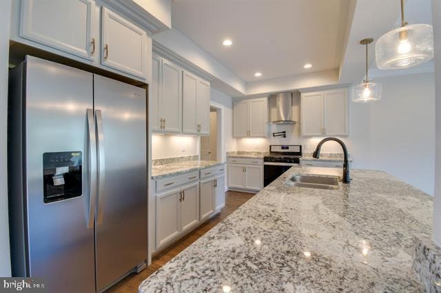 kitchen with sink, hanging light fixtures, stainless steel appliances, wall chimney range hood, and white cabinetry