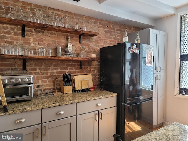 kitchen featuring brick wall, light stone counters, black refrigerator, and plenty of natural light