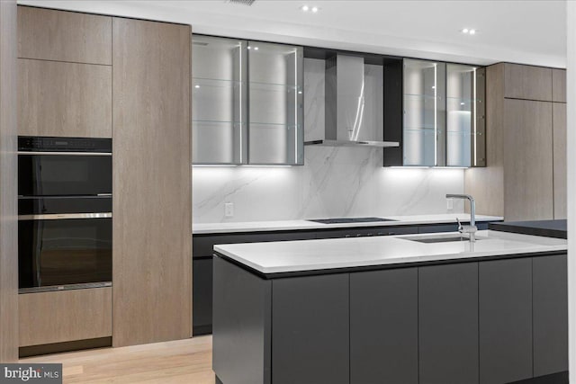 kitchen featuring wall chimney exhaust hood, cooktop, black double oven, sink, and light hardwood / wood-style flooring