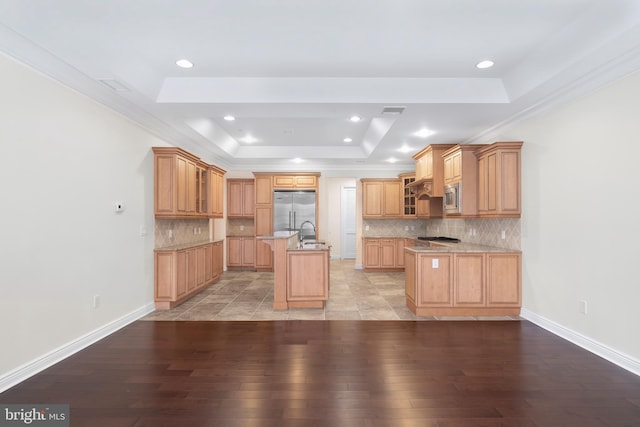 kitchen featuring an island with sink, built in appliances, light hardwood / wood-style floors, backsplash, and a raised ceiling