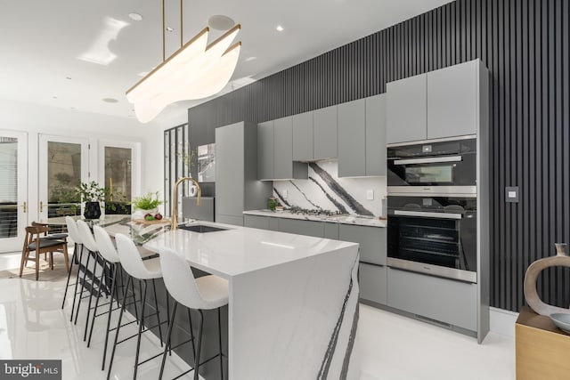 kitchen with decorative light fixtures, gray cabinets, a kitchen island with sink, double oven, and sink