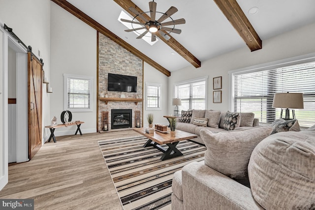 living room featuring ceiling fan, beamed ceiling, a barn door, a stone fireplace, and light wood-type flooring