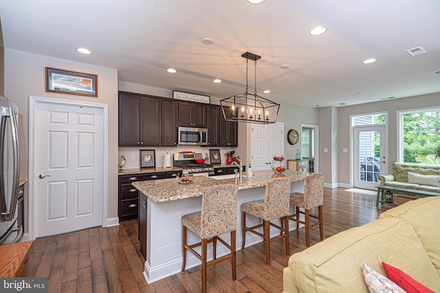 kitchen with pendant lighting, dark brown cabinets, an island with sink, appliances with stainless steel finishes, and dark hardwood / wood-style flooring