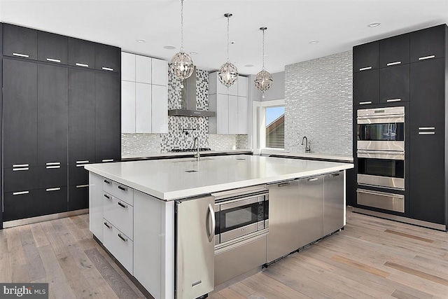 kitchen with a large island, stainless steel appliances, wall chimney range hood, pendant lighting, and light wood-type flooring