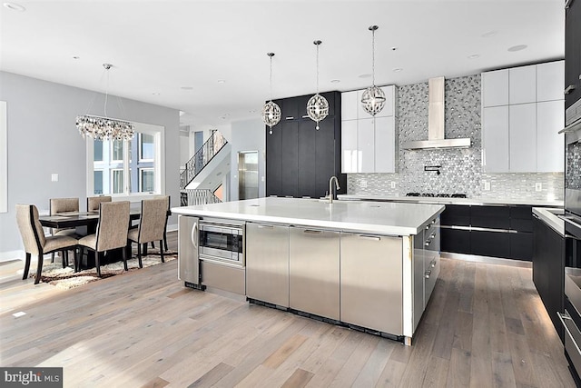 kitchen with wall chimney exhaust hood, light hardwood / wood-style floors, decorative light fixtures, a center island with sink, and appliances with stainless steel finishes