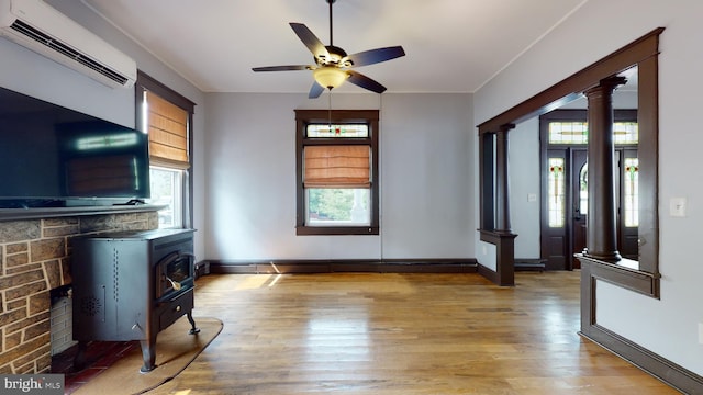 unfurnished living room with ornate columns, a wood stove, a wall mounted AC, light wood-type flooring, and ceiling fan