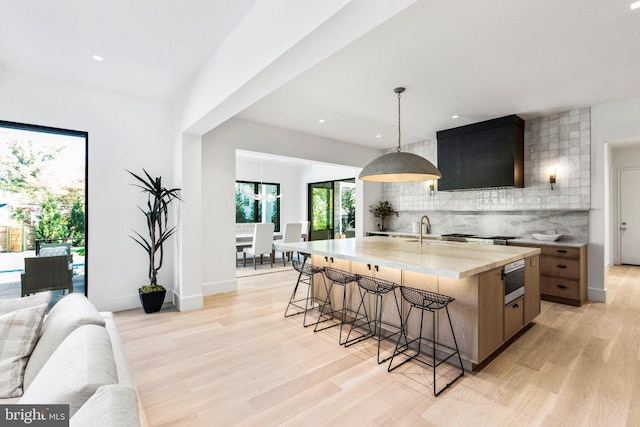 kitchen with tasteful backsplash, hanging light fixtures, a center island with sink, premium range hood, and light hardwood / wood-style flooring