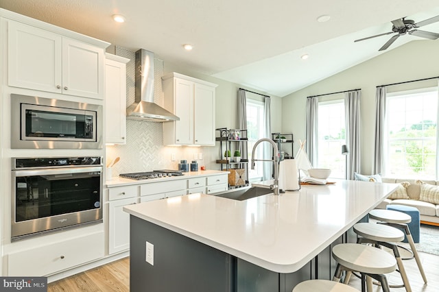 kitchen with wall chimney exhaust hood, a center island with sink, stainless steel appliances, and white cabinetry