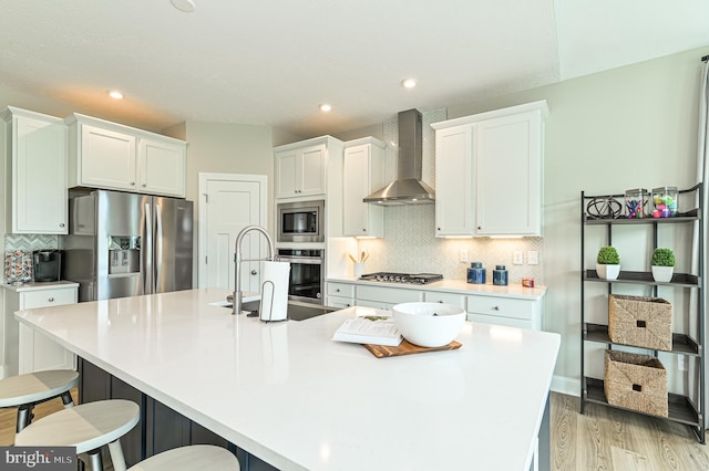 kitchen with white cabinetry, stainless steel appliances, a center island with sink, wall chimney range hood, and a breakfast bar area