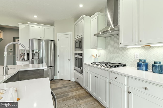 kitchen with appliances with stainless steel finishes, light wood-type flooring, tasteful backsplash, white cabinetry, and wall chimney exhaust hood