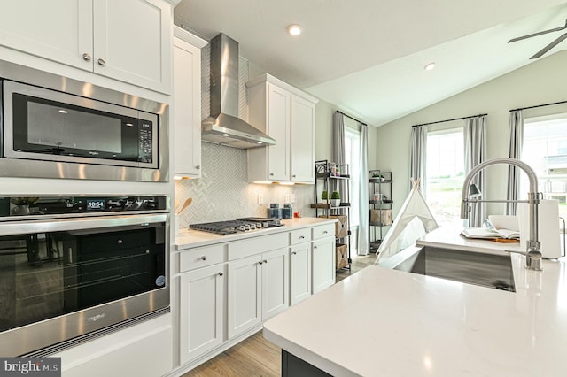 kitchen featuring tasteful backsplash, stainless steel appliances, light wood-type flooring, wall chimney exhaust hood, and ceiling fan