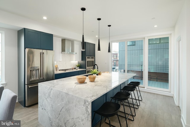 kitchen with a center island with sink, wall chimney exhaust hood, appliances with stainless steel finishes, hanging light fixtures, and light wood-type flooring