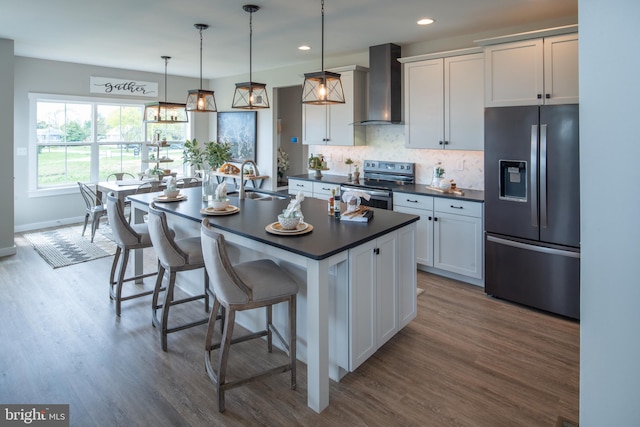 kitchen featuring appliances with stainless steel finishes, hanging light fixtures, an island with sink, white cabinets, and wall chimney range hood