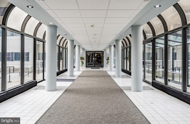 hallway featuring ornate columns, tile patterned floors, expansive windows, and a paneled ceiling
