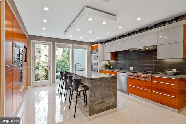 kitchen featuring appliances with stainless steel finishes, dark stone counters, an island with sink, light tile floors, and white cabinetry